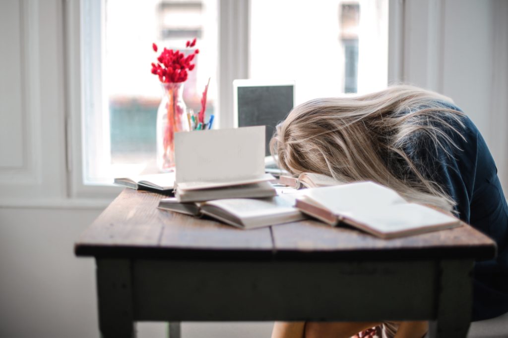 A woman is seen with her head resting on a table, surrounded by books and papers, suggesting exhaustion or burnout from studying or working.