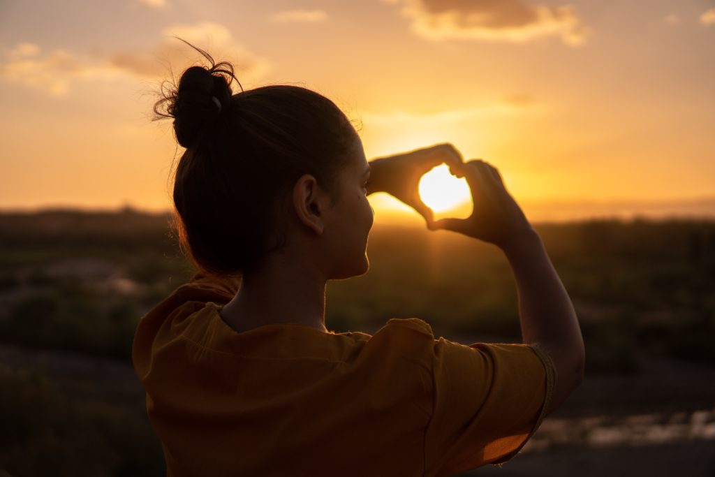 A woman stands on a hilltop with her arms outstretched, forming a heart shape with her hands as she gazes at the beautiful sunset sky with mountains in the background.